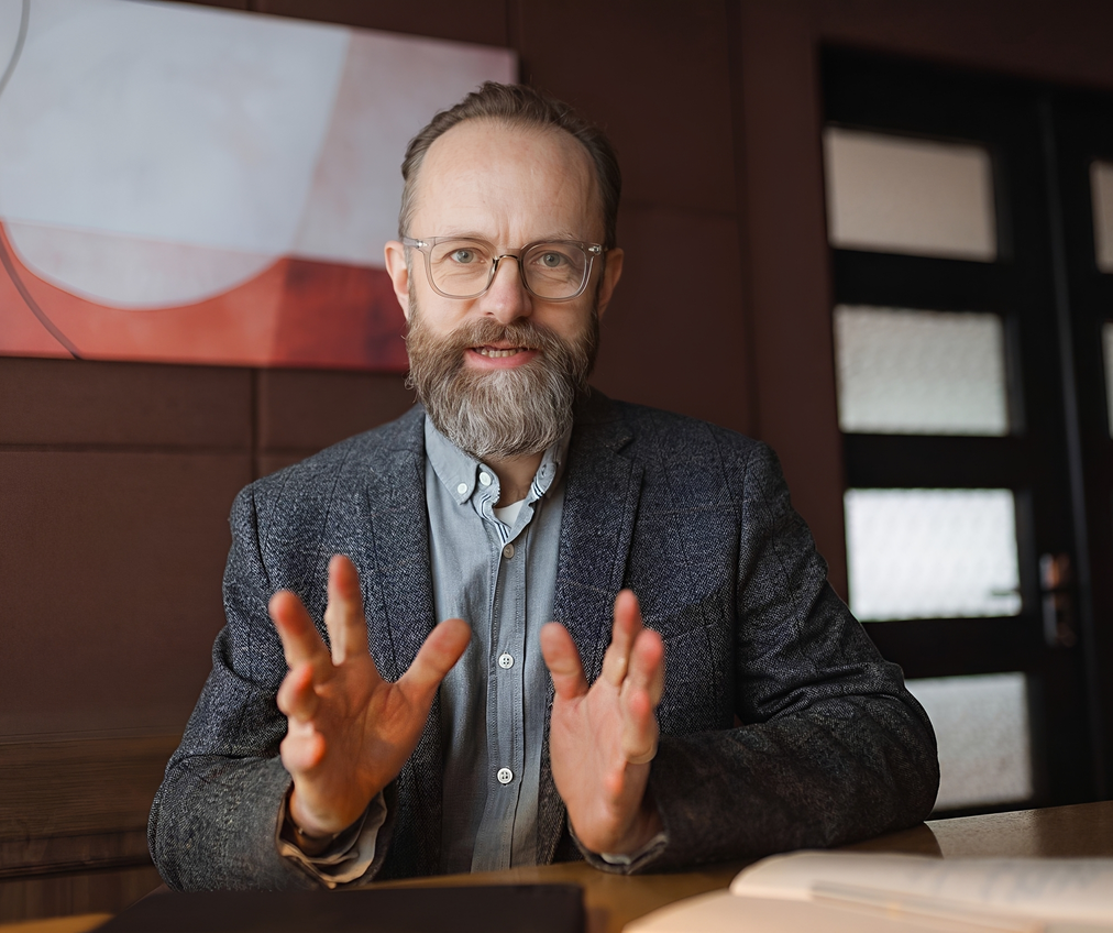 A professional middle-aged man with a graying beard, glasses, and a gray tweed blazer sitting at a wooden desk, speaking directly forward while gesturing with his hands open.