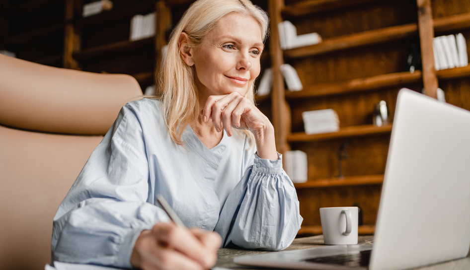 A blonde woman in a light blue blouse sits at a rustic wooden desk with a white coffee mug, smiling thoughtfully while looking at a laptop screen and writing with a pen.