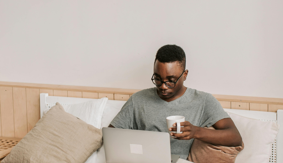 A young Black man wearing glasses sits cross-legged on a bed with pillows, holding a white coffee mug while looking down at an open silver laptop.