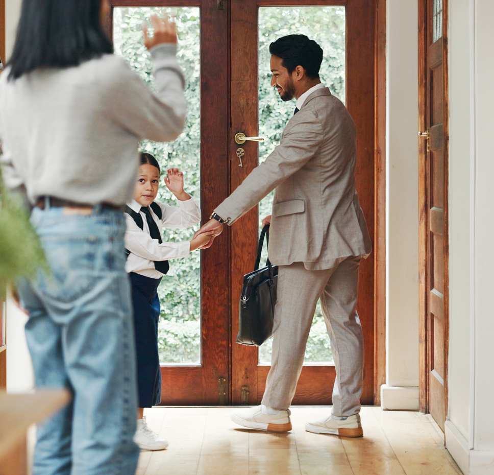 A man in a tan suit holding a briefcase stands by an open wooden front door, holding his young daughter's hand as she waves goodbye to a woman standing in the foreground inside a home.