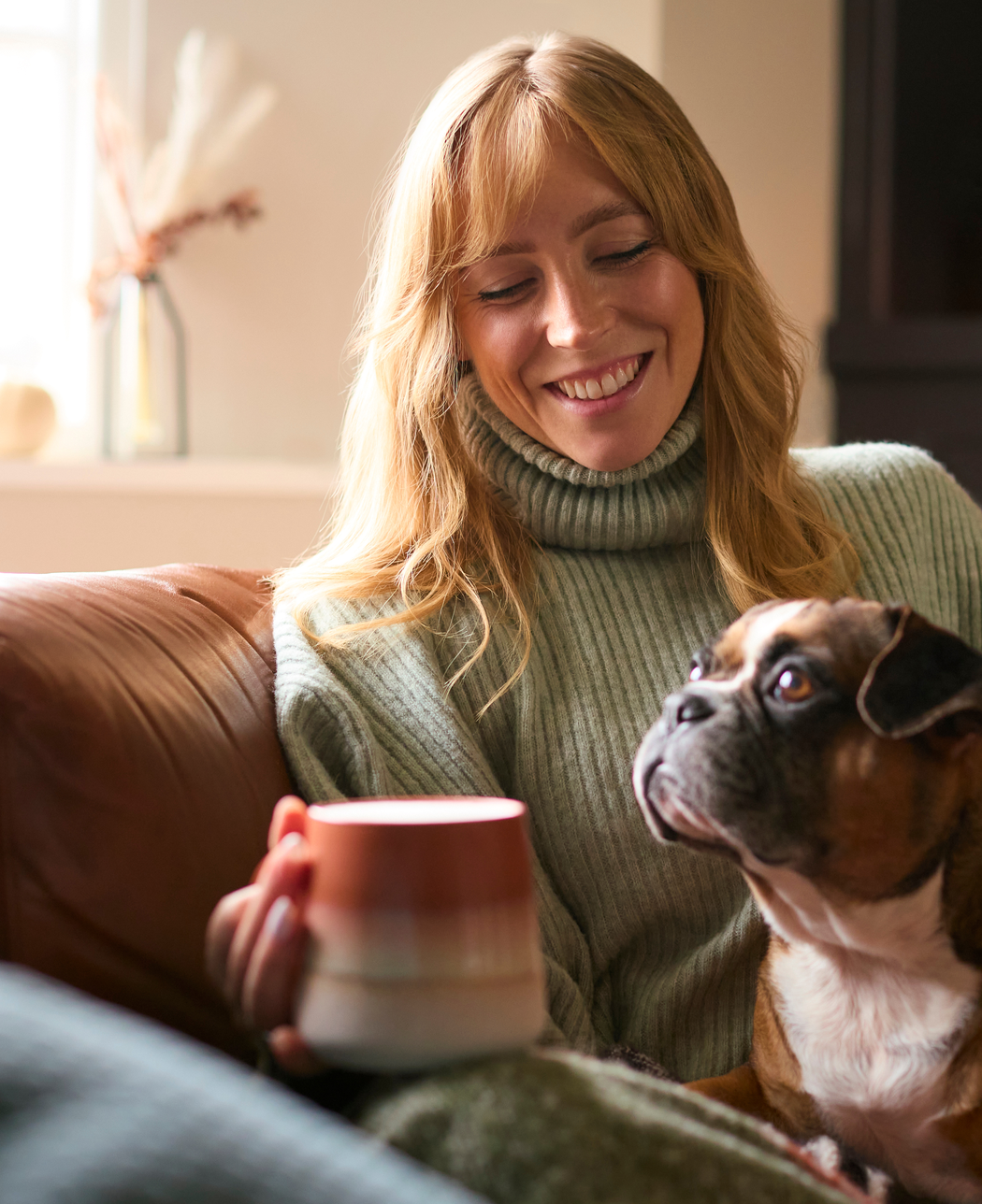 A smiling blonde woman wearing a light green knit turtleneck sweater sits on a brown leather couch, holding a colorful ceramic mug while looking warmly at her brown and white boxer dog.