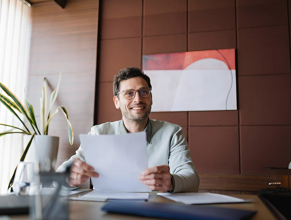 A smiling professional man with glasses sitting at an office desk, holding a white document and looking forward in a modern office with a potted plant.