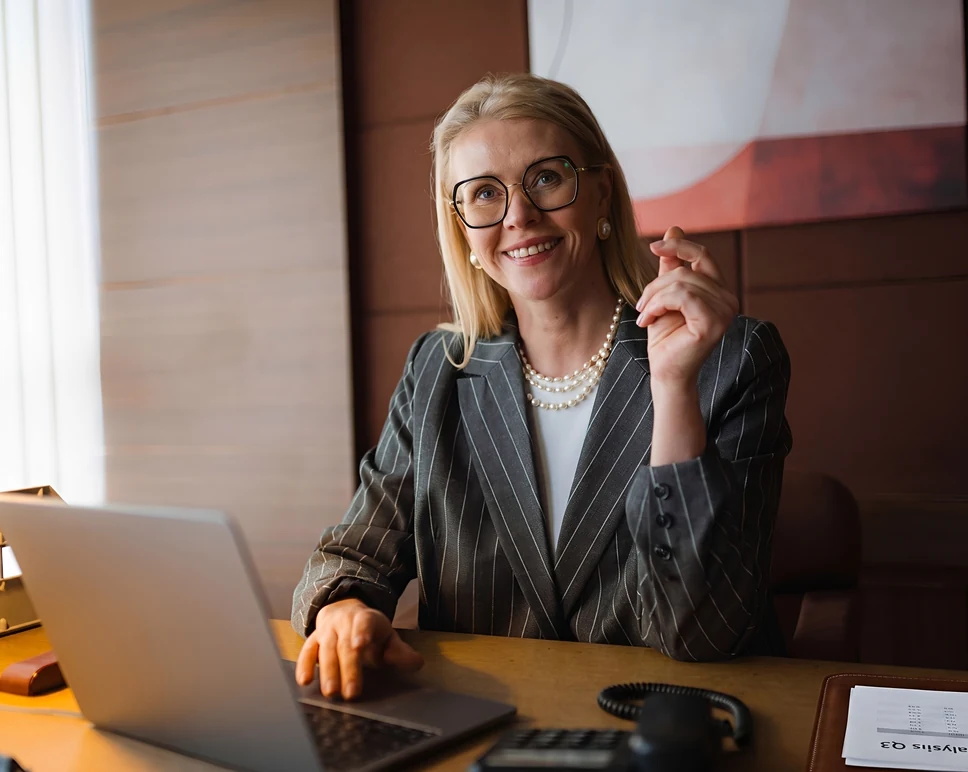 A professional blonde woman wearing glasses, a pinstripe blazer, and a pearl necklace smiling confidently while working on a laptop at an executive office desk.