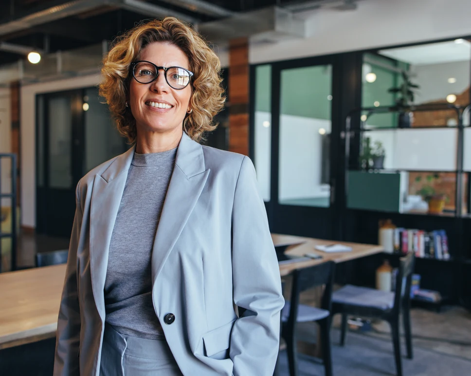 A confident female professional in a grey suit jacket and glasses smiling in a brightly lit, open-concept office background.