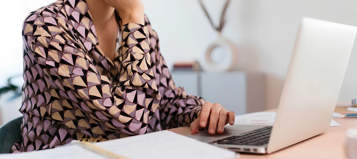 A close-up cropped view of a professional wearing a geometric patterned blouse sitting at a light wooden desk, with their hand resting on a silver laptop trackpad next to an open notebook.