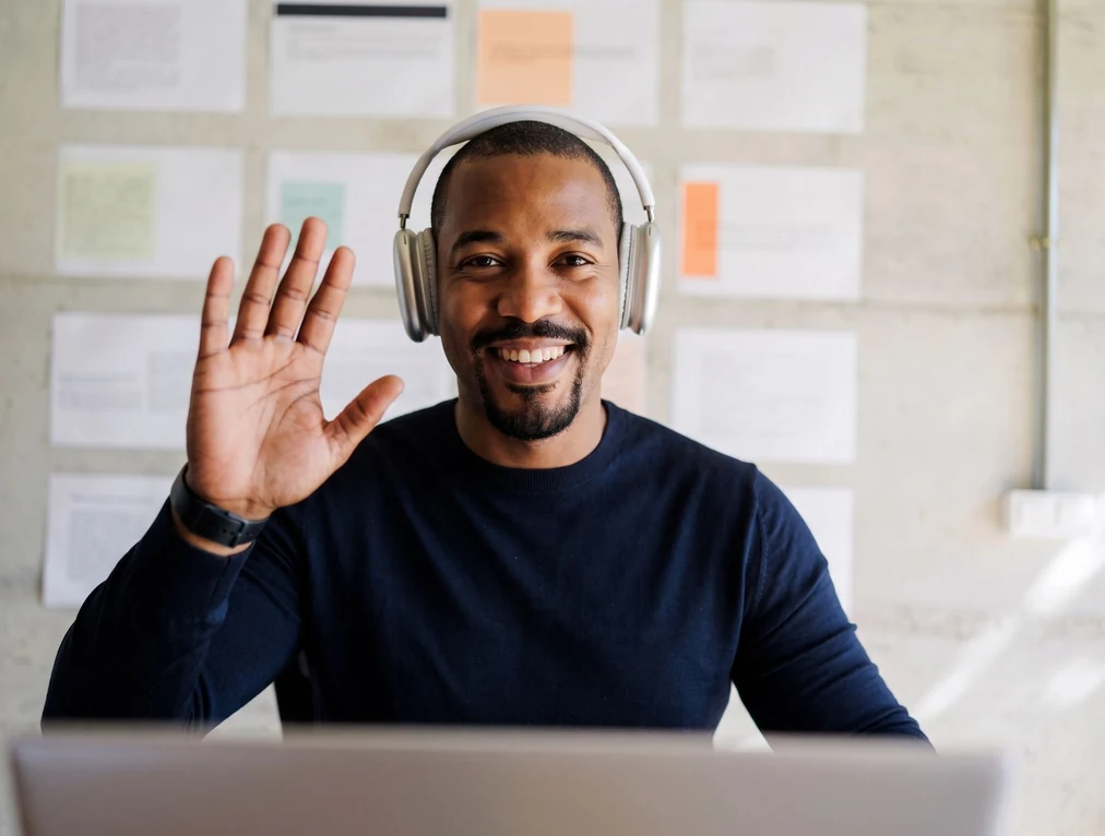 An approachable male professional in a dark long-sleeve shirt smiling and waving during a video conference call from his office desk.